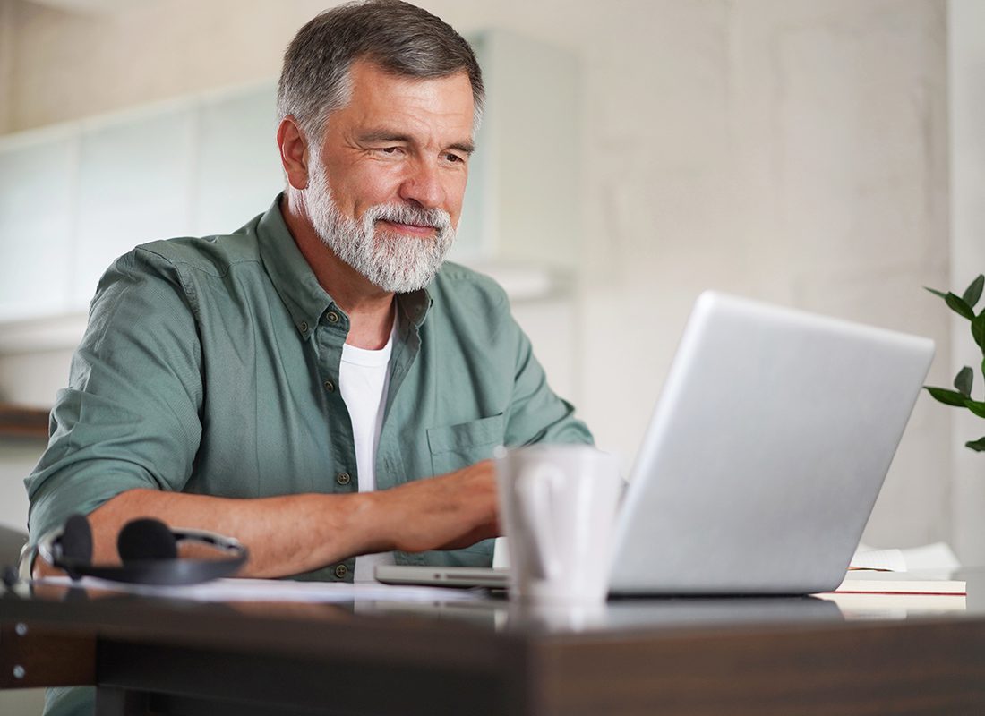 Blog - Older Man in Green Button Up Shirt Smiling While Staring at an Open Laptop and Sitting in his Home at a Table